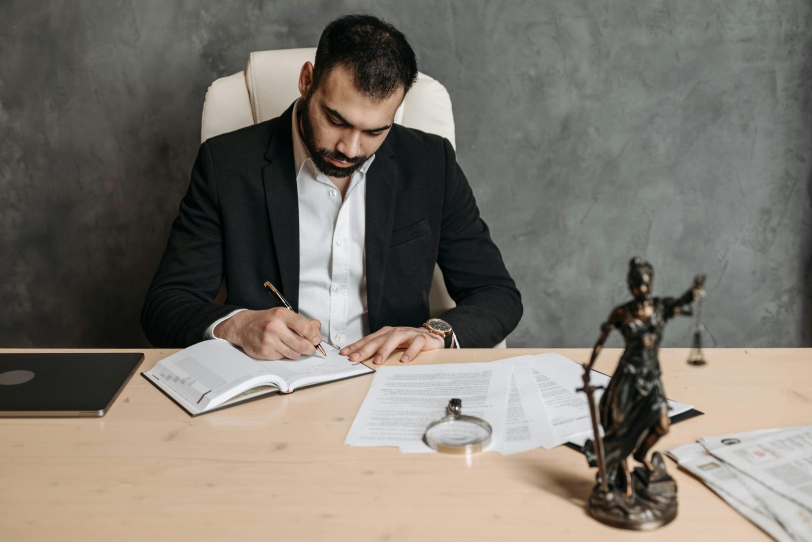 Focused lawyer in black suit at desk writing on documents in an office setting with legal statue.